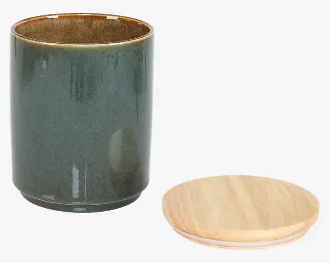 En cylindrisk beholder med en blank, mørkegrøn keramisk overflade og en glat, let afrundet kant. Indersiden har en brun, plettet finish. Beholderen leveres med et fladt, rundt låg af træ, der passer tæt på toppen og giver en lufttæt lukning til opbevaring.