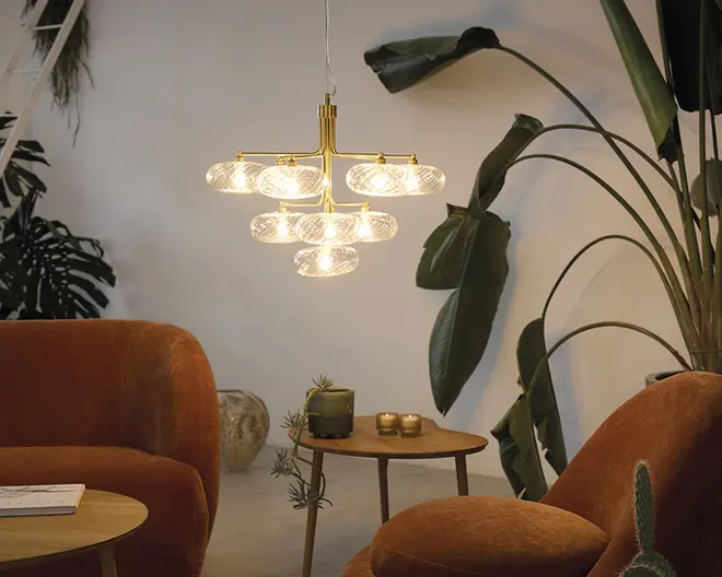 Billedet viser en moderne stue med en central pendellampe, der har flere klare, teksturerede glasglober arrangeret i to niveauer på en guldfarvet metalramme. Lampen udsender et varmt lys, der skaber en hyggelig stemning. Rummet indeholder rustfarvede fløjlslænestole, en matchende skammel og et rundt træsidebord med dekorative genstande såsom et stearinlys, en lille plante og guldfarvede tilbehør. Store grønne bladeplanter står i baggrunden og tilfører rummet et naturligt præg.