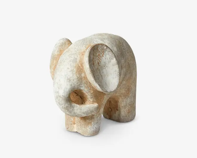 Håndskåret elefantfigur i træ med et rustikt, patineret finish. Designet er minimalistisk med bløde kurver og forenklede former, herunder et stort, rundt øre og en nedadbuet snabel. Overfladen har et slidt udseende med blandede lyse beige og hvide nuancer, hvilket giver figuren et antikt præg.