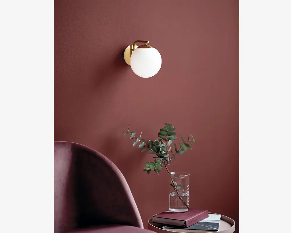 En vægmonteret lampe med en rund, mat hvid glasskærm og en base i guldfarvet metal. Lampen er monteret på en mat, mørkerødbrun væg. Under lampen står et lille rundt sidebord med en stak bøger og en klar glasvase med grønne eucalyptusgrene. Foran ses en del af en buet, polstret stol i samme mørkerødbrune farve.