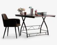 Et moderne arbejdsområde med et rektangulært skrivebord med lys træbordplade og sorte metalben i X-formet design. På bordet står en grøn skrivebordslampe, en vase med lyserøde blomster, en bærbar computer, et brunt krus, en stak bøger, en blyantholder med røde kuglepenne og en hvid skål med sort kant. Til skrivebordet hører en polstret stol med brun betræk, sorte armlæn og sorte, tilspidsede ben.