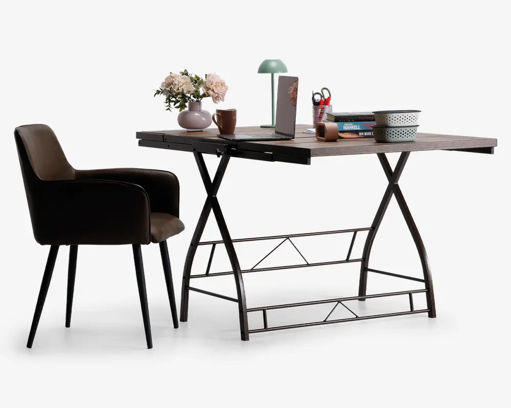 Et moderne arbejdsområde med et rektangulært skrivebord med lys træbordplade og sorte metalben i X-formet design. På bordet står en grøn skrivebordslampe, en vase med lyserøde blomster, en bærbar computer, et brunt krus, en stak bøger, en blyantholder med røde kuglepenne og en hvid skål med sort kant. Til skrivebordet hører en polstret stol med brun betræk, sorte armlæn og sorte, tilspidsede ben.