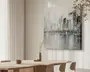 Billedet viser et moderne spiseområde med et bord og stole i lyst træ. På bordet står minimalistiske keramikvaser og skåle i neutrale farver, hvoraf én vase indeholder en enkelt tørret gren. På væggen hænger et stort, moderne maleri af en bysilhuet i afdæmpede grå, hvide og sorte nuancer, med en reflekterende vandoverflade nedenunder. Rummet er blødt oplyst af naturligt lys, der strømmer ind gennem lette gardiner, hvilket skaber en rolig og elegant stemning.