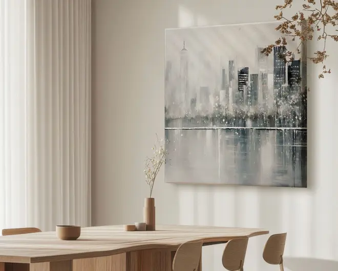 Billedet viser et moderne spiseområde med et bord og stole i lyst træ. På bordet står minimalistiske keramikvaser og skåle i neutrale farver, hvoraf én vase indeholder en enkelt tørret gren. På væggen hænger et stort, moderne maleri af en bysilhuet i afdæmpede grå, hvide og sorte nuancer, med en reflekterende vandoverflade nedenunder. Rummet er blødt oplyst af naturligt lys, der strømmer ind gennem lette gardiner, hvilket skaber en rolig og elegant stemning.