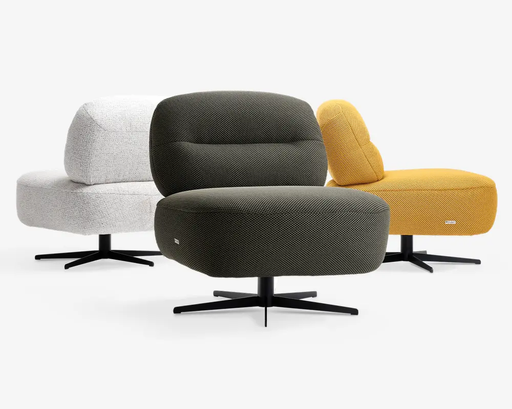 Tre moderne loungestole med afrundede, polstrede sæder og rygstøtter, alle betrukket med tekstureret stof. Stolene vises i tre farver: lysegrå, olivengrøn og sennepsgul. Hver stol har en sort, firebenet drejefod, og designet lægger vægt på komfort med brede, bløde siddepladser og en let tilbagelænet ryg.