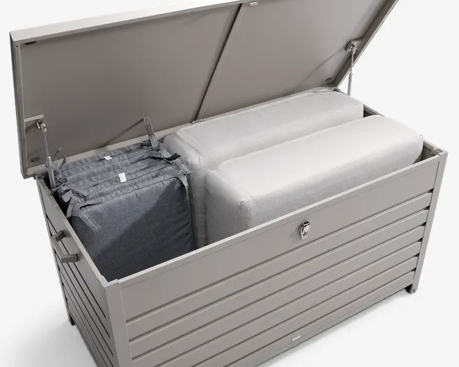 Stor, rektangulær udendørs opbevaringsboks med hængslet låg, vist åben for at vise det rummelige indre. Boksen er lavet af lysegråt metal med horisontale lameller for et moderne udseende. Indeni er der to store, lyse hynder og én mørkegrå hynde, hvilket viser den store opbevaringskapacitet. Låget har en lås for sikkerhed og gasdæmpere for nem åbning og lukning.