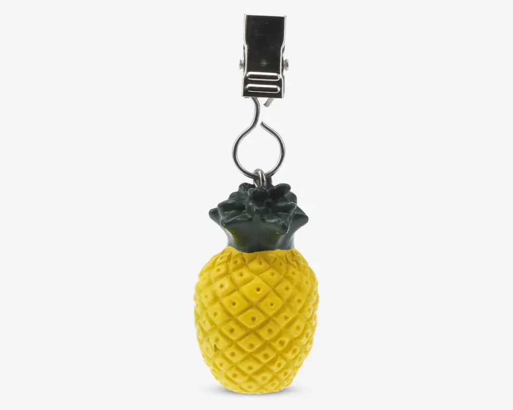 Et dekorativt element formet som en ananas med en gul, mønstret overflade og grønne blade i toppen. Den er fastgjort til en sølvfarvet metalclips, hvilket gør den velegnet til ophængning eller fastgørelse på andre genstande.