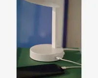 Nærbillede af en moderne hvid lampe med rund fod, der har en indbygget USB-opladerport. Et hvidt opladerkabel er tilsluttet porten, og en smartphone er forbundet og ligger i nærheden. Lampen har et minimalistisk design og står på en grøn overflade.