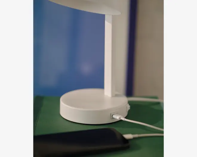 Nærbillede af en moderne hvid lampe med rund fod, der har en indbygget USB-opladerport. Et hvidt opladerkabel er tilsluttet porten, og en smartphone er forbundet og ligger i nærheden. Lampen har et minimalistisk design og står på en grøn overflade.