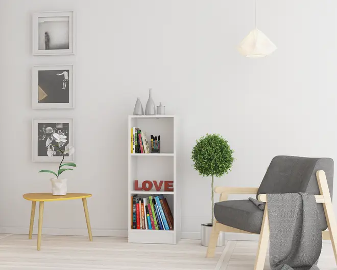 Et moderne, minimalistisk hjørne af en stue med en lænestol i lyst træ, mørkegrå hynder og et matchende tæppe lagt over armlænet. Ved siden af stolen står et lille, rundt gult sidebord med skrå ben, hvorpå der står en hvid potteplante. En høj, hvid reol står op ad væggen og viser en blanding af bøger, dekorative vaser og et rødt 'LOVE'-skilt. Ved siden af reolen står en rund, grøn topiary-plante i en sølvfarvet potte. Væggen er dekoreret med tre sort-hvide indrammede kunsttryk, der er arrangeret lodret. Rummet er godt oplyst med en hvid pendellampe, og farvepaletten er overvejende neutral med farveindslag fra bordet og bøgerne.