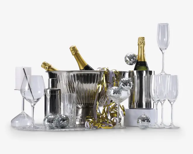 Et festligt arrangement med to flasker champagne i sølvfarvede isspande, omgivet af forskellige typer glas såsom vinglas, champagneglas og tumblere. Opsætningen inkluderer også en cocktailshaker, metalliske guldfarvede bånd og flere små sølvfarvede diskokugle-dekorationer, hvilket skaber en elegant og festlig stemning.