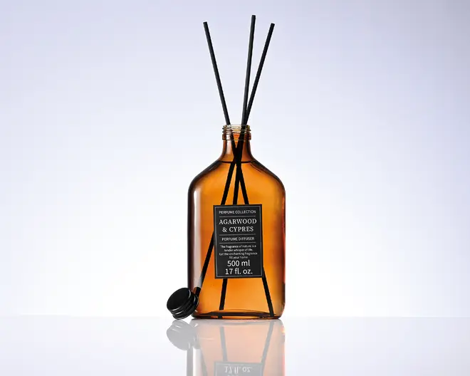 Amberfarvet glasflaske med tre sorte duftpinde og et sort etiketmærke med teksten "Agarwood & Cypress", 500 ml, samt en sort prop ved siden af.