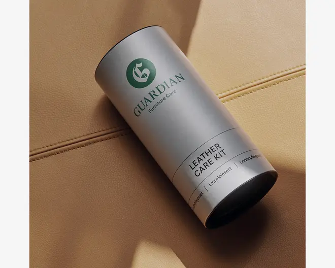 En cylindrisk beholder med mat sølvfinish, der har et grønt logo og mærket 'Guardian Furniture Care' trykt på forsiden. Beholderen er mærket 'Leather Care Kit' og har en sort bund. Emballagen fremstår robust og moderne, designet til opbevaring af produkter til læderpleje. Den er fotograferet på en lysebrun læderoverflade med synlige syninger, hvilket antyder dens anvendelse til vedligeholdelse af læder.