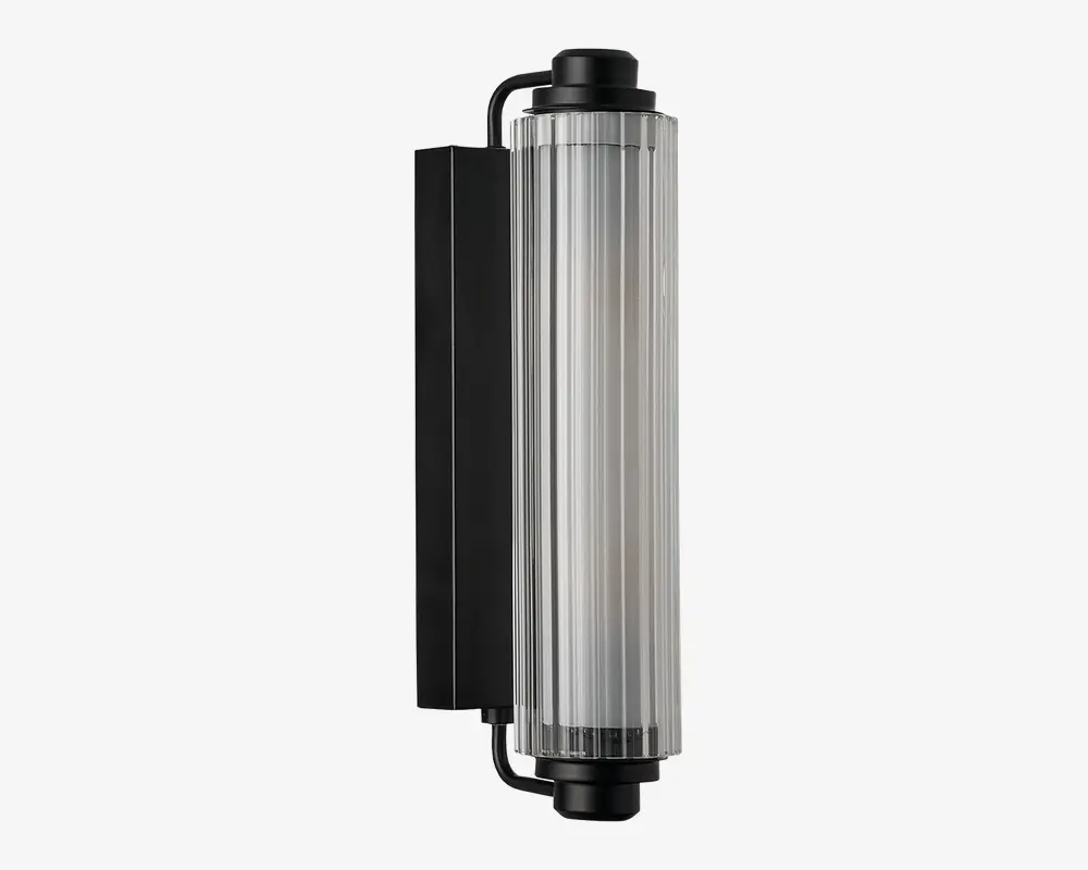Væghængt lampe med en lodret cylindrisk glasskærm med rillet struktur, fastholdt af en mat sort metalramme. Designet er moderne og industrielt med rene linjer og en solid, rektangulær bagplade. Glasset giver et diffust lys, hvilket gør lampen velegnet til både indendørs og udendørs brug.