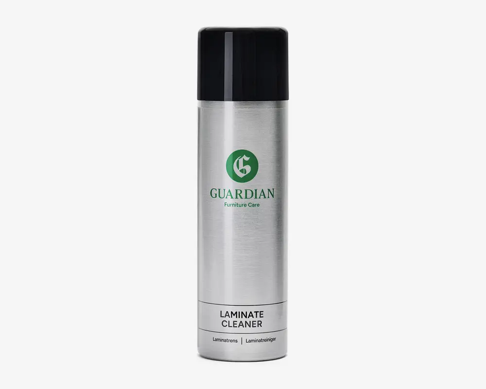 Høj, cylindrisk sølvfarvet spraydåse med sort låg. Forsiden har et grønt logo og teksten 'Guardian Furniture Care'. Nederst på dåsen står produktnavnet 'Laminate Cleaner' med fed skrift samt oversættelser på andre sprog. Designet er stilrent og moderne med en metallisk overflade.