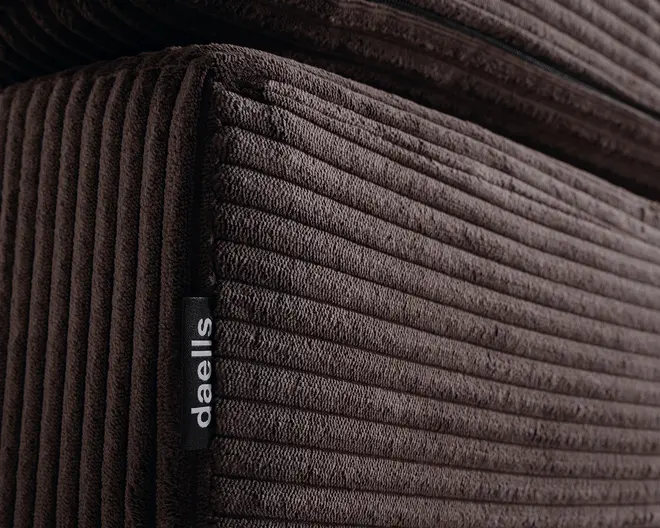 Nærbillede af mørkebrunt fløjl med ribbet struktur, der viser detaljeret syning og en blød, plysset overflade. Et lille sort stofmærke med mærket 'daells' er syet ind i sømmen, hvilket angiver mærket. Kanterne er pænt afsluttede, og materialet virker tykt og slidstærkt, velegnet til komfortabel sidde- eller liggeplads.