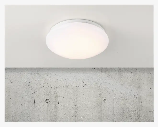 En rund, loftmonteret lampe med en glat, hvid, matteret skærm, der fordeler lyset jævnt. Lampen har et minimalistisk design med lav profil, hvilket gør den velegnet til moderne indretning. Lampen ser ud til at være lavet af plast eller glas og er monteret på et ensfarvet, hvidt loft.