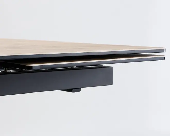 Nærbillede af en bordplade med lys træfinish og et slankt, moderne udtryk. Bordet har en sort metalramme og en integreret udtræksplade eller hylde under hovedfladen, hvilket giver ekstra arbejdsplads eller opbevaring. Designet er minimalistisk og nutidigt med rene linjer og en glat overflade.