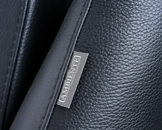 Nærbillede af en sort læderoverflade med tydelig struktur og fine syninger langs kanten. En lille, rektangulær stofmærkat er syet ind i sømmen og viser mærkenavnet 'AMBIENTE' samt teksten 'ITALIAN DESIGN' med hvid skrift. Læderet fremstår som værende af høj kvalitet og slidstærkt, velegnet til eksklusive møbler eller tilbehør.