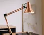 Justerbar skrivebordslampe med en metallisk kobberfinish, der har en konisk lampeskærm og dobbelte fjederbelastede arme for fleksibel placering. Lampen har et slankt, moderne design med sorte og sølvfarvede beslag ved leddene, hvilket gør det nemt at justere vinklen. Indersiden af skærmen er hvid for optimal lysrefleksion.
