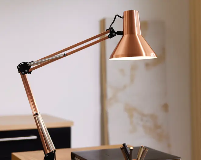 Justerbar skrivebordslampe med en metallisk kobberfinish, der har en konisk lampeskærm og dobbelte fjederbelastede arme for fleksibel placering. Lampen har et slankt, moderne design med sorte og sølvfarvede beslag ved leddene, hvilket gør det nemt at justere vinklen. Indersiden af skærmen er hvid for optimal lysrefleksion.