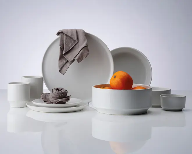 Et sæt minimalistisk, mat hvidt keramikstel bestående af store og små tallerkener, skåle og krus. Delene har rene, moderne linjer og en glat overflade. En af skålene indeholder flere appelsiner, hvilket tilføjer et farverigt element. En grå stofserviet er lagt over en tallerken, og en anden serviet er rullet sammen og placeret på en tallerken, hvilket antyder en borddækning. Udtrykket er elegant og moderne.
