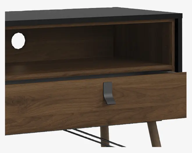 Nærbillede af et møbel med mat sort top, en åben hylde med et rundt hul til kabelgennemføring bagtil og en nederste skuffe i træ med et moderne metalhåndtag. Træet har en naturlig årestruktur, og benene er skråtstillede, hvilket giver et retro-inspireret udtryk.