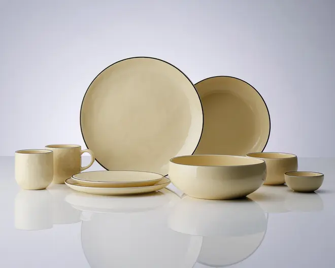 Et sæt cremefarvet keramikservice med blank overflade og en diskret mørk kant langs kanterne. Sættet indeholder store og små tallerkener, skåle i forskellige størrelser samt krus med behagelige hanke. Designet er minimalistisk og moderne med bløde, afrundede former og ensartet farve.