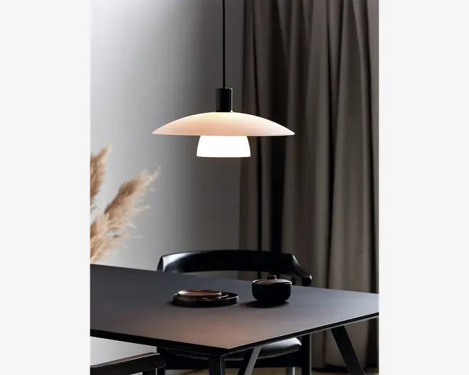 En moderne pendellampe med en bred, lav, mat hvid skærm og en mindre cylindrisk diffuser nedenunder, ophængt i en sort ledning. Lampen udsender et blødt, varmt lys, der skaber en hyggelig stemning over et minimalistisk sort spisebord. Bordet er dækket med en sort skål og en matchende tallerken, og en sort stol er delvist synlig. I baggrunden ses gardiner i neutrale farver og dekorativ pampasgræs.
