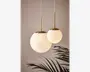 To hængelamper med kugleformede, matthvide glasskærme og detaljer i børstet messing. Den ene kugle er større og hænger lidt lavere end den mindre. Begge lamper er ophængt i hvide ledninger. Designet er moderne og elegant og giver et blødt, varmt lys. En bladplante ses delvist nederst i billedet.