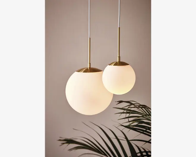 To hængelamper med kugleformede, matthvide glasskærme og detaljer i børstet messing. Den ene kugle er større og hænger lidt lavere end den mindre. Begge lamper er ophængt i hvide ledninger. Designet er moderne og elegant og giver et blødt, varmt lys. En bladplante ses delvist nederst i billedet.