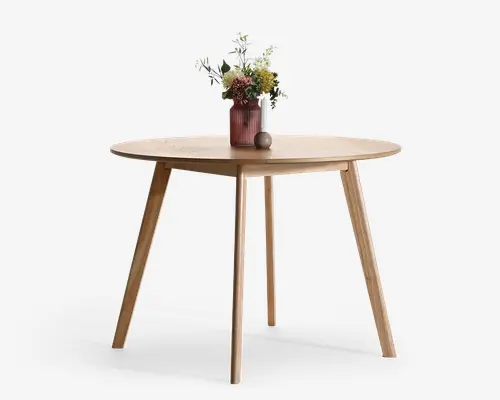 Rundt bord i lyst træ med glat bordplade og fire tilspidsede ben, vist med en lille vase med tørrede blomster og en lille dekorativ kugle ovenpå.
