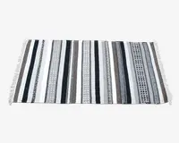 Rektangulært tekstil med skiftende lodrette striber i sorte, hvide, grå og brune nuancer. Striberne har forskellige mønstre, herunder ensfarvede, geometriske former og prikdesigns. Begge korte sider er afsluttet med hvide frynser.