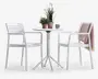 Et moderne udendørs cafésæt bestående af to hvide stole med armlæn og lamel-ryg samt et rundt hvidt bord med firebenet søjlebase. Bordet er pyntet med to potteplanter og et blåt krus. Den ene stol har et lyserødt strikket tæppe hængende over ryglænet. Sættet har et rent, minimalistisk design, der passer til terrasser eller altaner.