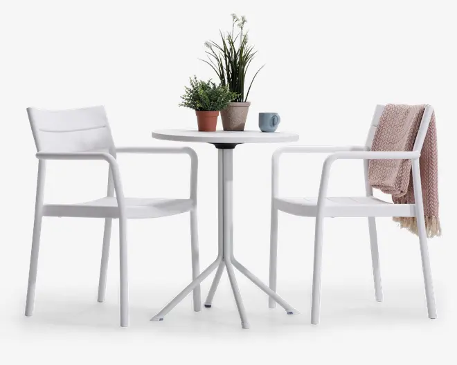 Et moderne udendørs cafésæt bestående af to hvide stole med armlæn og lamel-ryg samt et rundt hvidt bord med firebenet søjlebase. Bordet er pyntet med to potteplanter og et blåt krus. Den ene stol har et lyserødt strikket tæppe hængende over ryglænet. Sættet har et rent, minimalistisk design, der passer til terrasser eller altaner.