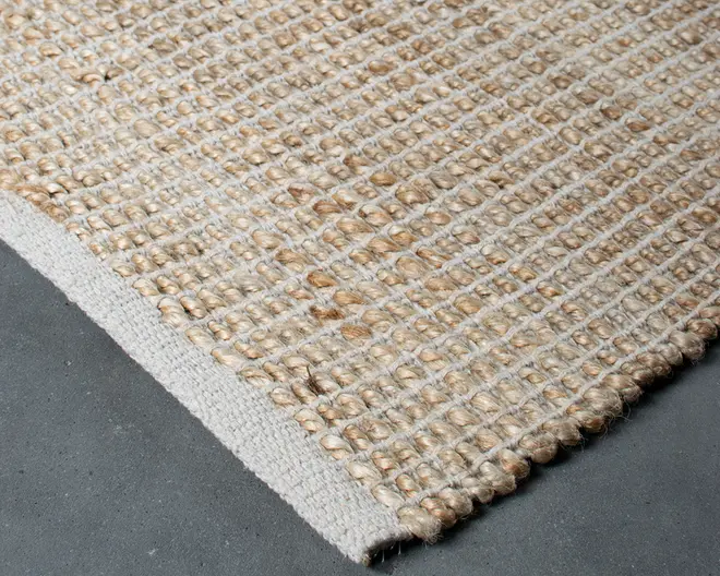 Nærbillede af et vævet tæppe med en kraftig, tekstureret overflade lavet af naturfibre i beige og lysebrune nuancer. Vævemønstret er tæt og ensartet, hvilket giver et gitterlignende udseende. Kanten er pænt afsluttet med en solid, lysere kant for ekstra holdbarhed og et rent udtryk. Tæppet virker tykt og robust, velegnet til områder med meget trafik.