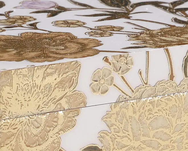 Nærbillede af en æske med blank hvid overflade, dekoreret med indviklede blomster-mønstre i guld og beige. Blomsterne er præget, hvilket giver et tekstureret og luksuriøst udseende. Designet har store blomster og mindre blomster med fine detaljer på kronblade og stilke.