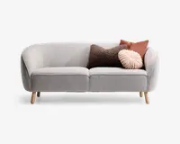 En moderne, lysegrå sofa med buet ryg og armlæn, betrukket med stof og stående på fire lyse træben. Sofaen er pyntet med tre dekorative puder: en firkantet brun pude med tekstur, en firkantet rustfarvet pude og en rund, plisseret pudderrosa pude, som giver et hyggeligt og stilfuldt udtryk.