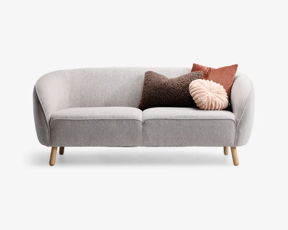 En moderne, lysegrå sofa med buet ryg og armlæn, betrukket med stof og stående på fire lyse træben. Sofaen er pyntet med tre dekorative puder: en firkantet brun pude med tekstur, en firkantet rustfarvet pude og en rund, plisseret pudderrosa pude, som giver et hyggeligt og stilfuldt udtryk.