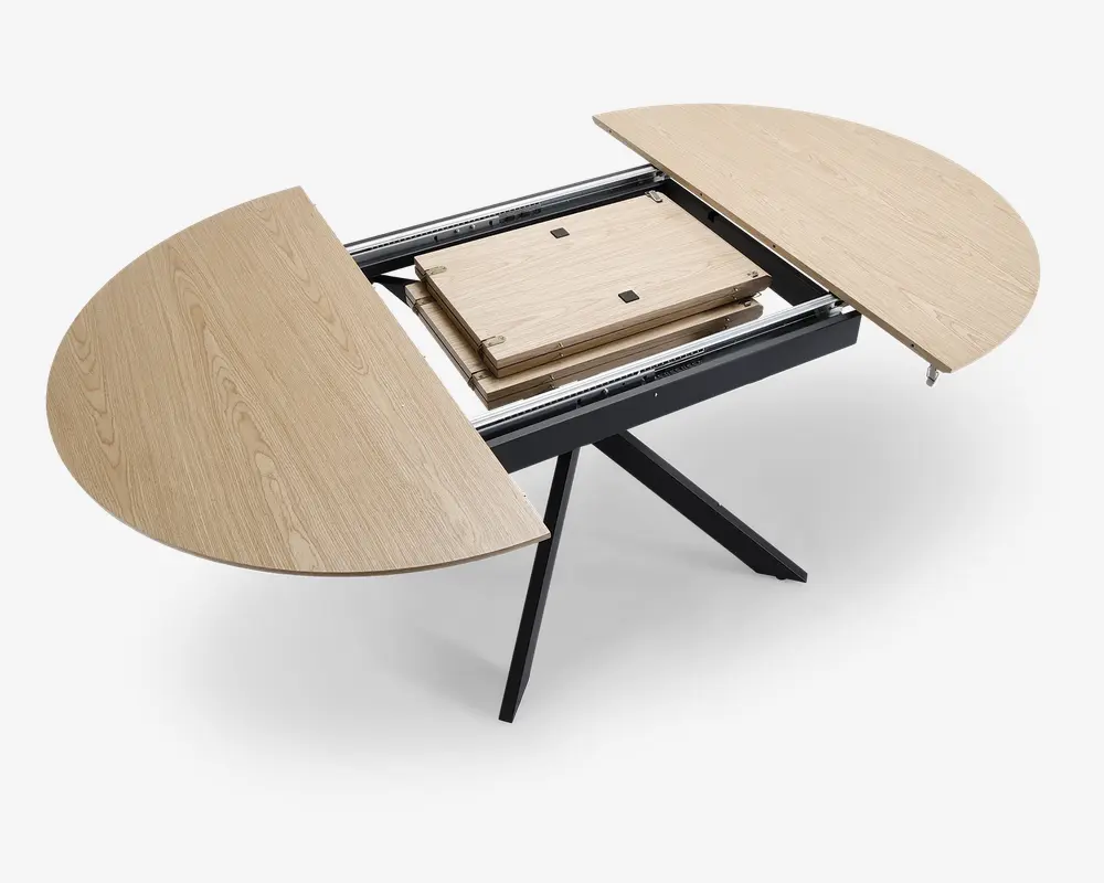 Billedet viser et bord i lyst træ med en rund bordplade, der er delt i to halvdele og åbnet op for at vise et udtræksmekanisme i midten. Bordet har en skjult tillægsplade, som opbevares i midten og kan trækkes ud for at udvide bordets overflade. Bordpladen har en naturlig træstruktur, og bordet står på skrå sorte metalben for stabilitet.