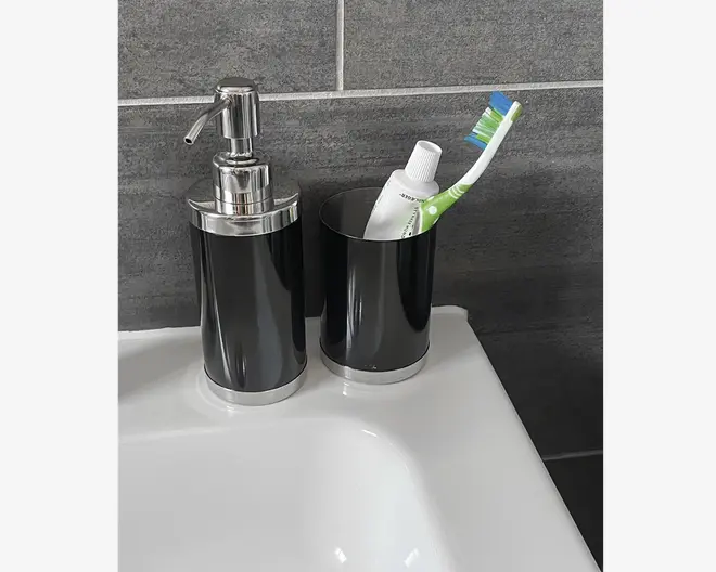 Et sæt matchende badeværelsestilbehør i blank sort med sølvfarvede detaljer. Sættet består af en sæbedispenser med pumpe og et krus, der bruges til at holde en tandbørste og tandpasta. Designet er moderne og stilrent med glatte, cylindriske former og en reflekterende overflade. Produkterne står på en hvid håndvask foran en mørkegrå flisevæg.