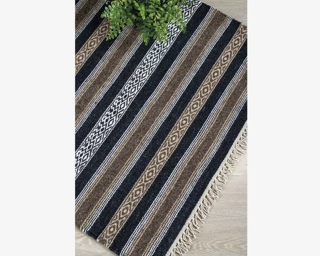 Rektangulært vævet tekstil med skiftevis sorte, brune og hvide striber samt geometriske diamantmønstre. Kanterne er afsluttet med korte, cremefarvede frynser. Materialet ser tekstureret og slidstærkt ud, velegnet til brug på gulvet. En grøn plante er delvist synlig i øverste venstre hjørne for at give en fornemmelse af størrelsen.