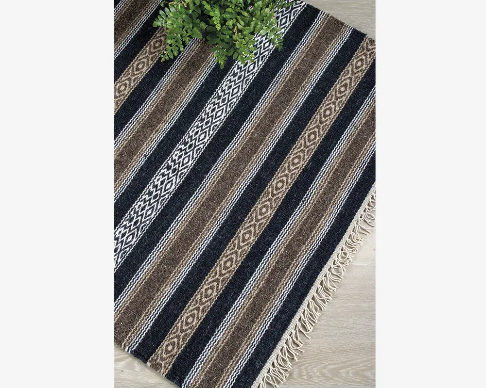 Rektangulært vævet tekstil med skiftevis sorte, brune og hvide striber samt geometriske diamantmønstre. Kanterne er afsluttet med korte, cremefarvede frynser. Materialet ser tekstureret og slidstærkt ud, velegnet til brug på gulvet. En grøn plante er delvist synlig i øverste venstre hjørne for at give en fornemmelse af størrelsen.