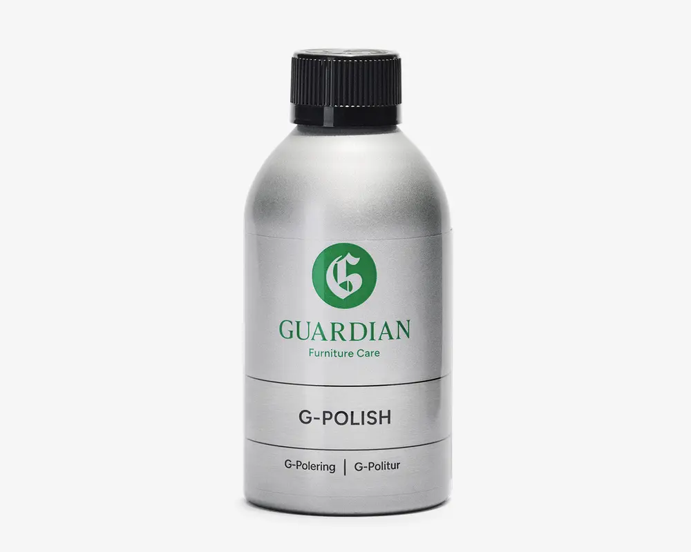 En sølvfarvet flaske med sort skruelåg, der har et grønt 'G'-logo og teksten 'GUARDIAN Furniture Care' på etiketten. Produktet er mærket 'G-POLISH' og har yderligere tekst på flere sprog. Flasken har et stilrent, metallisk udseende og et enkelt, moderne design.