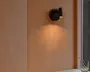 Mattsort væglampe med rund base og kort, justerbart cylindrisk lampehoved, der kaster et varmt lys nedad på en væg med træpaneler.