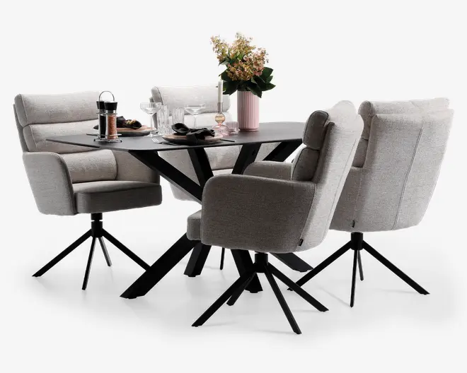 Billedet viser et moderne spiseområde med et rektangulært bord med mat sort overflade og vinklede sorte metalben. Rundt om bordet står fire polstrede stole i lysegråt stof, hver med polstrede armlæn og ryglæn for ekstra komfort. Stolene har sorte metalstel med drejefunktion og fire ben. Bordet er dækket med glas, tallerkener, bestik og en stempelkande, og der står en lyserød vase med en blomsterbuket som dekoration.