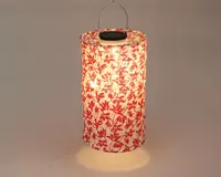 Cylindrisk lanterne med hvid baggrund og rødt blomstermønster. Lanternen er oplyst indefra og giver et varmt skær. Den har et metalhåndtag øverst til ophængning, og materialet ser let ud, muligvis papir eller stof.