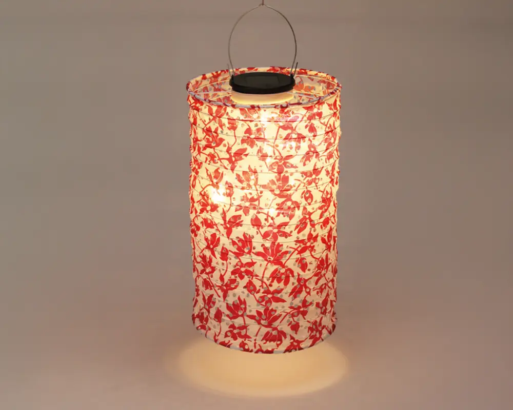 Cylindrisk lanterne med hvid baggrund og rødt blomstermønster. Lanternen er oplyst indefra og giver et varmt skær. Den har et metalhåndtag øverst til ophængning, og materialet ser let ud, muligvis papir eller stof.