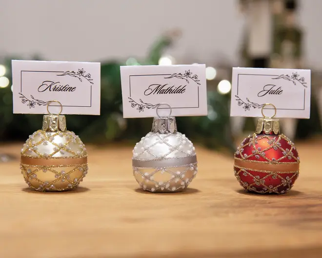 Tre dekorative julekugler bruges som bordkortsholdere. Hver kugle har en metallisk overflade – én i guld, én i sølv og én i rød – med glitrende krydsmønstre og et vandret bånd omkring midten. Kuglerne har en metalring i toppen, som holder et rektangulært bordkort med elegant skrift og blomsterkant. Kortene har forskellige navne. Kuglerne står på en træoverflade med en sløret, festlig baggrund.
