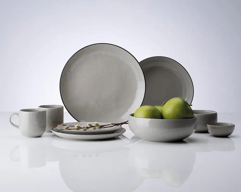 Et sæt keramisk service med et minimalistisk design, blank lysegrå overflade og diskrete mørkere kanter. Sættet indeholder store og små tallerkener, skåle, krus og en lille skål. Delene har en glat, let afrundet form og et moderne, enkelt udtryk. Skålen er vist med grønne æbler, hvilket fremhæver dens rummelige størrelse.